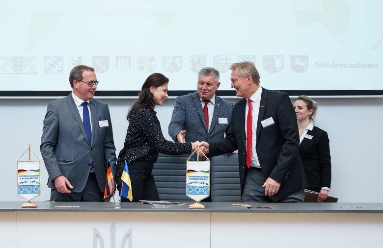 Feierlicher Handschlag zwischen einer Frau und einem Mann. Zwei weitere Männer und eine Frau stehen im Hintergrund. Im Vordergrund sind Symbole der deutsch-ukrainischen Zusammenarbeit zu sehen.
